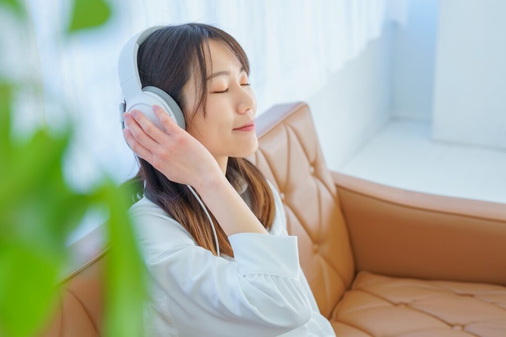 A woman sitting on a sofa with her eyes closed, listening to music through white headphones. She has a gentle smile, creating a calm and relaxed atmosphere : Body Scan Meditation