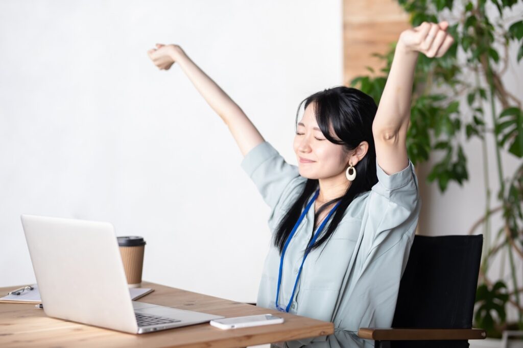A woman sitting in front of a laptop in the office, stretching her arms with a relaxed expression. She closes her eyes, looking refreshed and comfortable : Body Scan Meditation