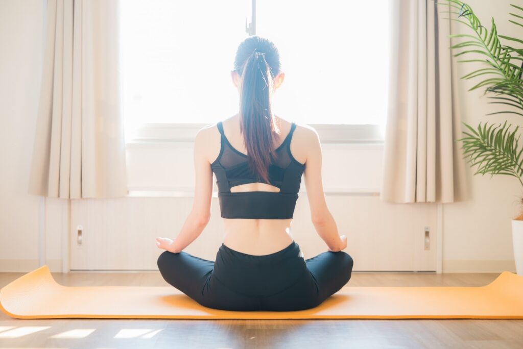 明るい部屋で、ヨガマットの上に座って瞑想する女性。黒いスポーツウェアを着て、背筋を伸ばし、窓からの光に包まれて静かに座っている:ガイド付き筋弛緩法