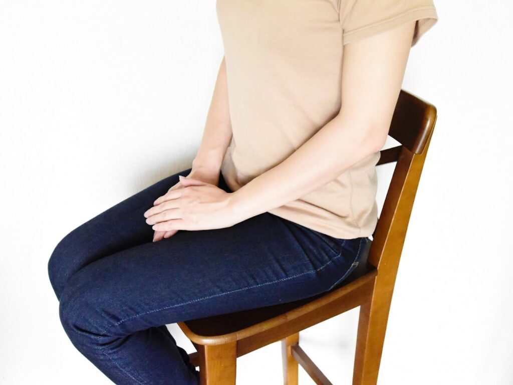 椅子に座り、両手を膝の上にそっと重ねている人の上半身。ベージュのTシャツとジーンズを着ており、落ち着いた姿勢で座っている:ガイド付き筋弛緩法