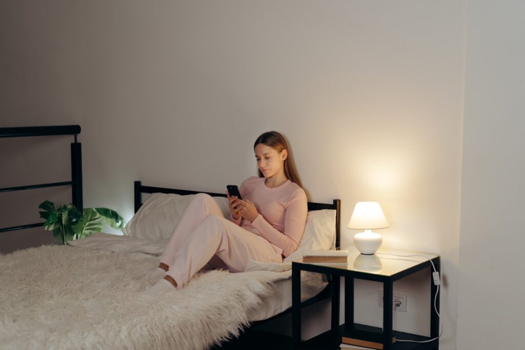 ベッドの上でピンクのルームウェアを着た女性がスマートフォンを見ている。枕元には小さなテーブルと白いランプ、本が置かれ、柔らかな照明が部屋を温かく照らしている:ガイド付き筋弛緩法