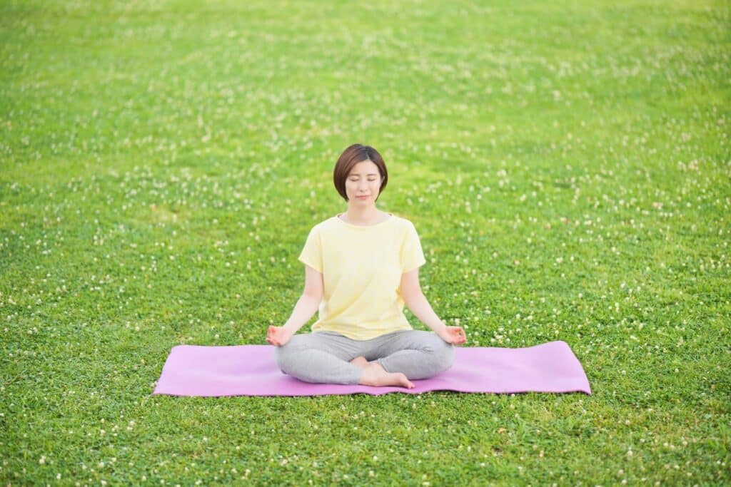 緑の芝生の上で、女性がヨガマットに座り目を閉じて瞑想している。黄色いTシャツとグレーのパンツを着て、穏やかな朝の空気を感じながらリラックスしている：Matter Over Mind