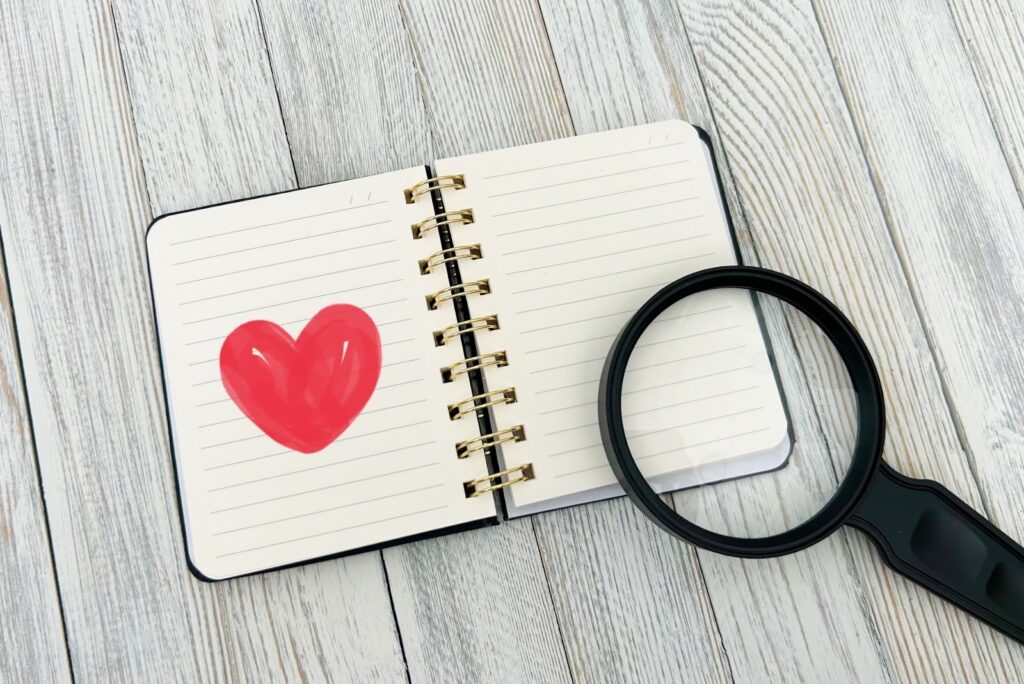 木のテーブルの上に開かれたノートと虫眼鏡。ノートの左ページには赤いハートが描かれており、愛や感謝、気づきをテーマにしたイメージ：Matter Over Mind
