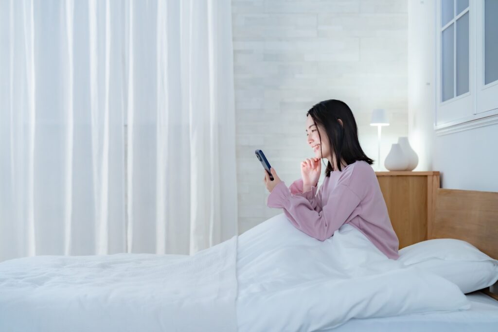 ベッドに座った女性がスマートフォンを見ながら笑顔を浮かべている。白いカーテンとランプがある明るい寝室の風景:睡眠催眠