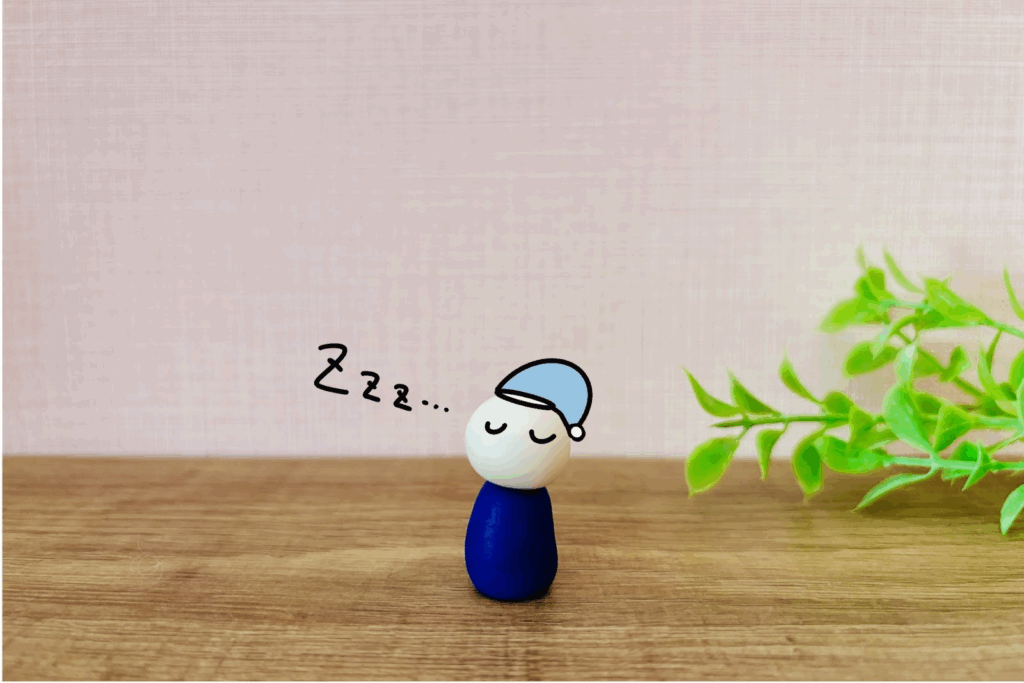 A wooden doll with a sleepy expression wearing a blue nightcap, shown with "Zzz…" as if sleeping, with green leaves placed beside it :Sleep Meditation