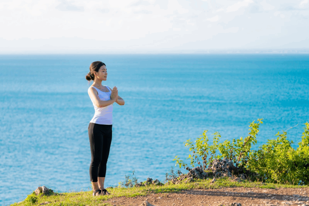 A woman wearing a white tank top and black pants stands with her palms together in a meditative pose by the sea, surrounded by the calm blue ocean and sky, embodying serenity and mindfulness :Meditation Positions