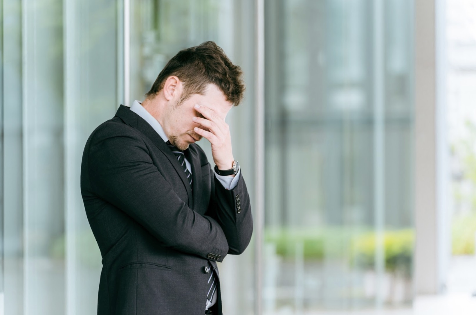 A man in a suit standing outdoors with his hand on his forehead, expressing stress, frustration, or worry: MBSR