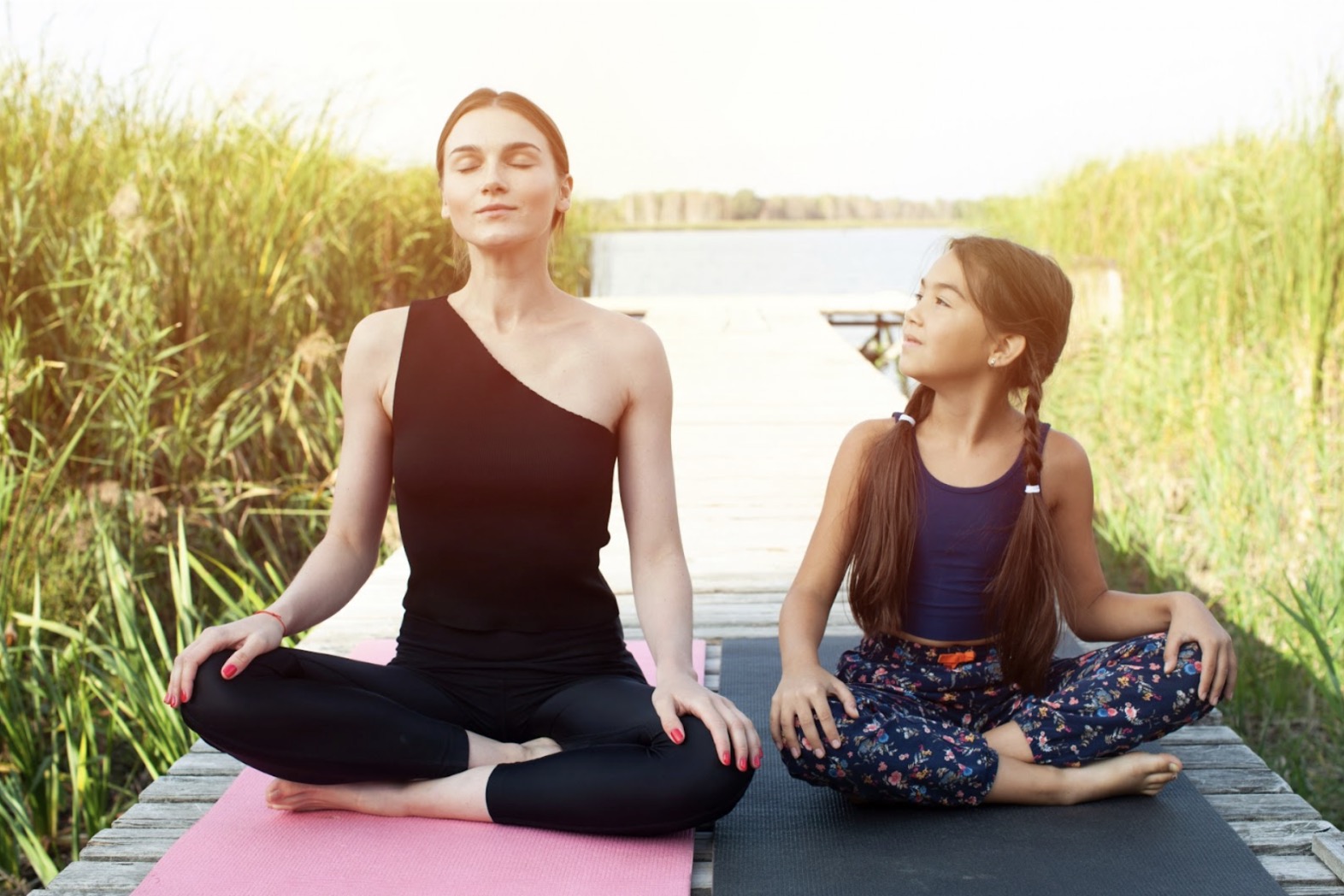 湖のほとりでヨガマットに座り、瞑想する母親とその様子を見つめる娘。自然の中で穏やかな時間を過ごしている：瞑想の方法