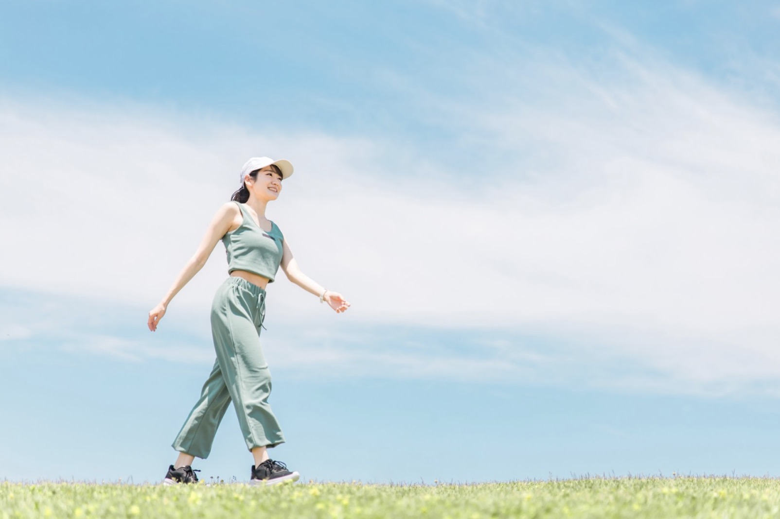 青空の下、芝生の上を笑顔で歩く女性。キャップとスポーツウェアを身につけ、健康的で爽やかな印象を与える：マインドフルネス・アクティビティ