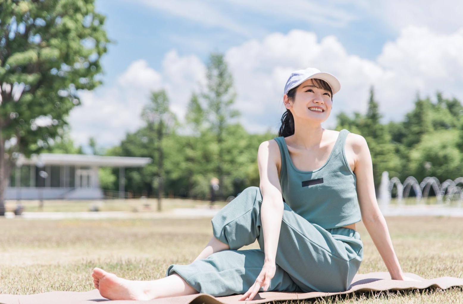 公園の芝生の上でヨガマットに座り、笑顔でストレッチをする女性。青空と木々に囲まれ、心地よい陽射しの中でリラックスした表情を見せている：マインドフルネス・エクササイズ