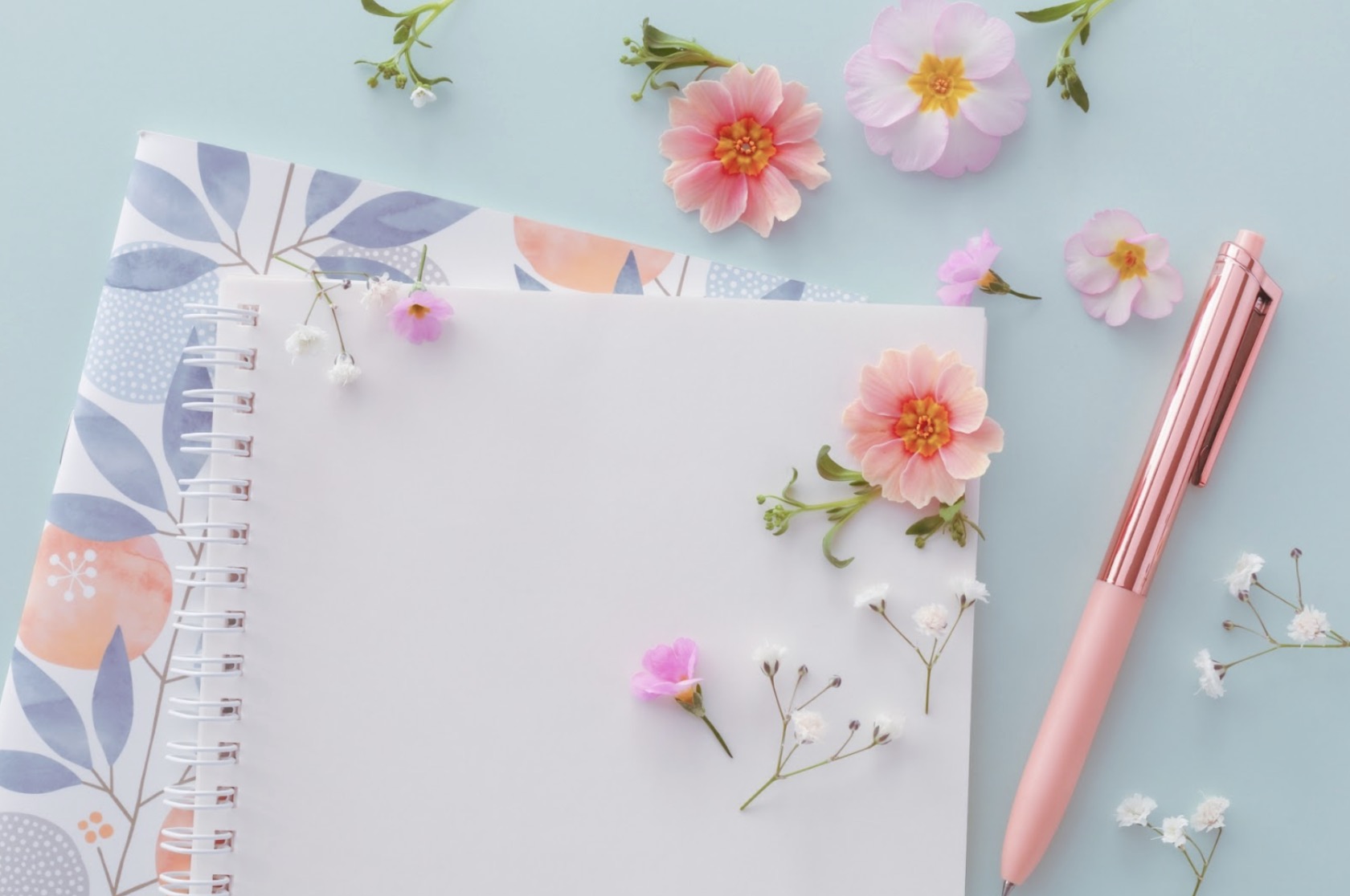 A pastel-toned flat lay with flowers scattered around a notebook and a pink pen, evoking a bright and gentle spring atmosphere for journaling or writing: Mindfulness Journal