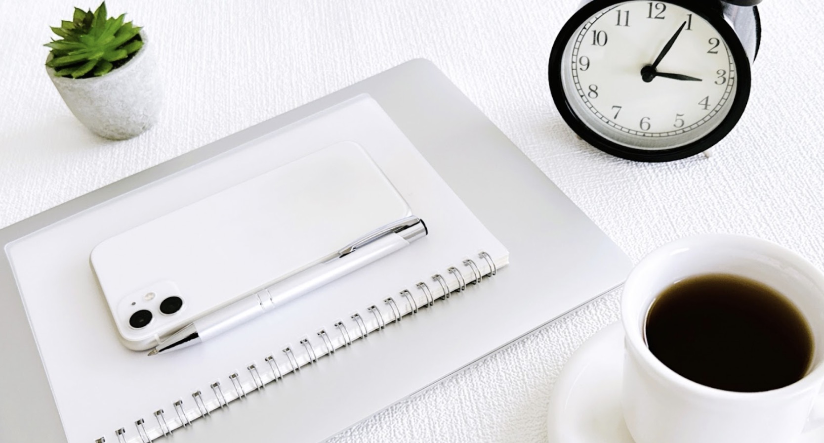 A minimalist workspace with a laptop, notebook, smartphone, pen, coffee cup, clock, and a small succulent plant neatly arranged on a white desk: Mindfulness Techniques
