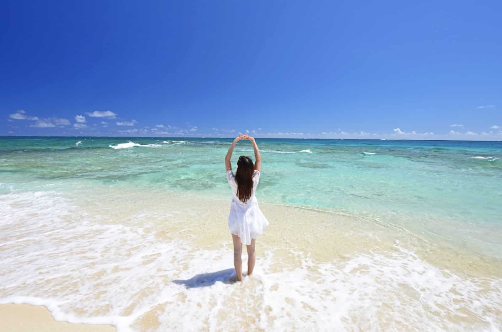 A woman in a white dress stands at the edge of a clear turquoise sea, raising her arms toward the bright blue sky. The scene captures a sense of freedom and the refreshing beauty of summer: Relaxation Technique