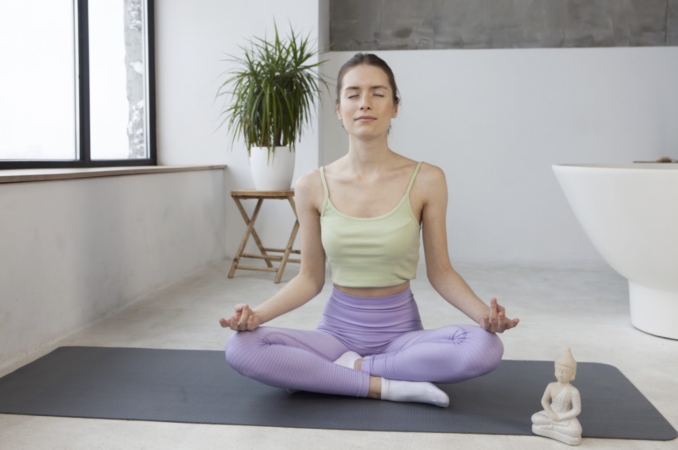 A woman meditating cross-legged on a yoga mat, wearing a light green top and lavender leggings, with a small Buddha statue beside her in a bright and calm indoor space: TM Meditation