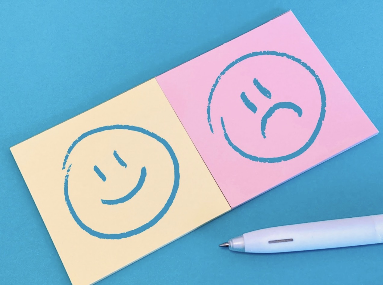 A yellow sticky note with a smiley face and a pink sticky note with a sad face placed side by side on a blue background, with a white pen nearby — symbolizing contrasting emotions: The Chimp Paradox