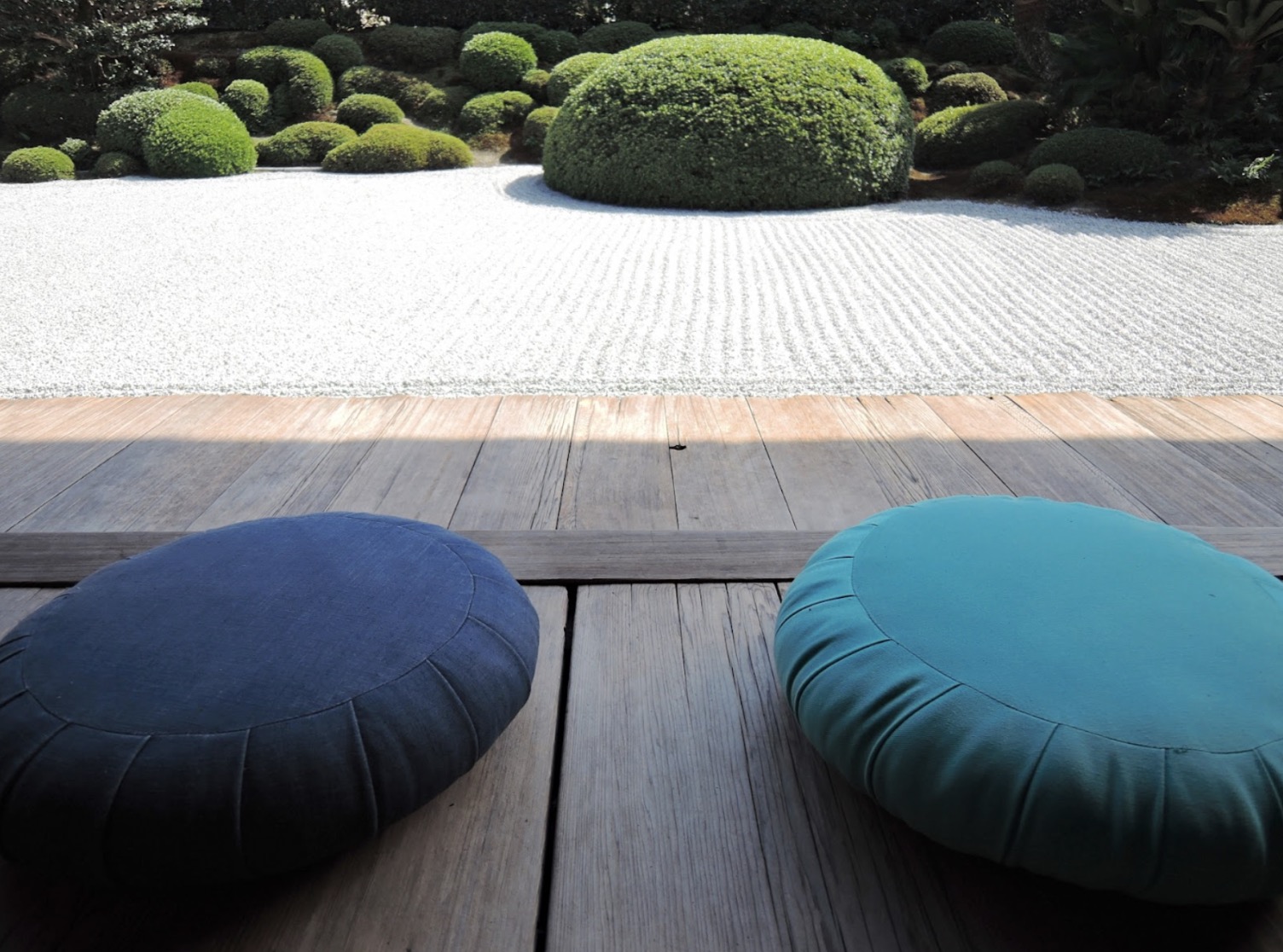 Two round cushions placed on a wooden veranda overlooking a Japanese rock garden with white gravel and neatly trimmed green bushes, evoking tranquility and harmony: Zafu