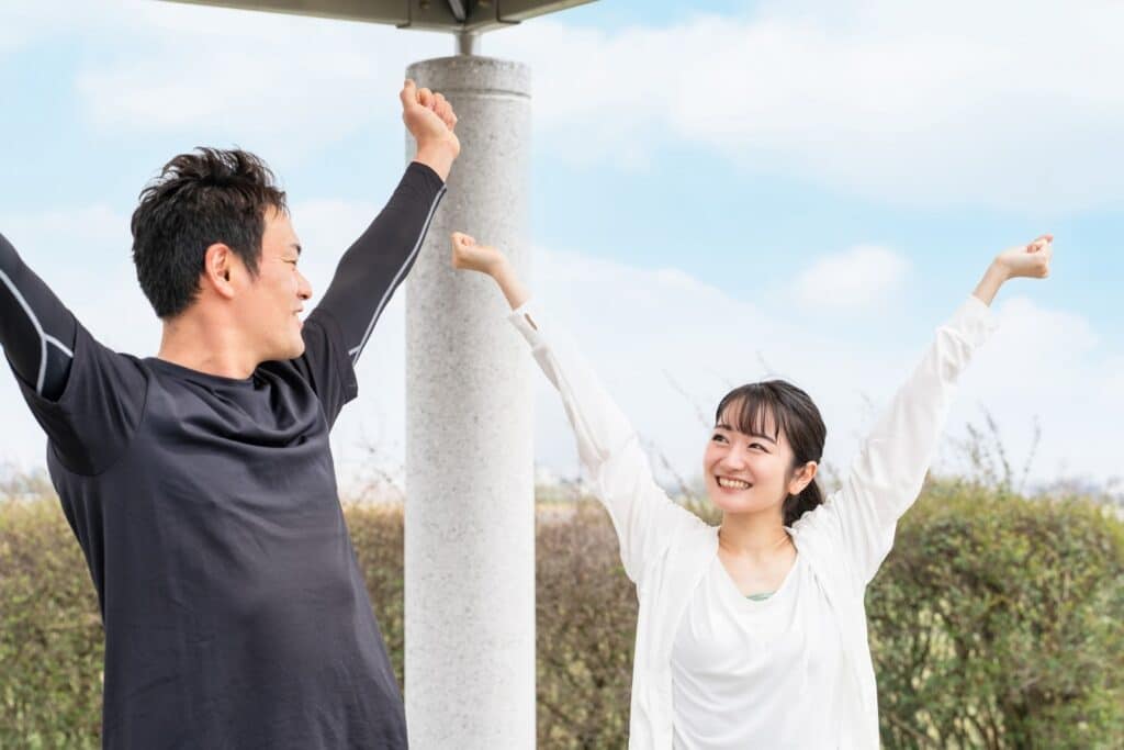 屋外でストレッチをしている男女。青空の下でお互いに笑顔を交わしながら両手を伸ばし、爽やかな朝の運動を楽しんでいる：リラックス法