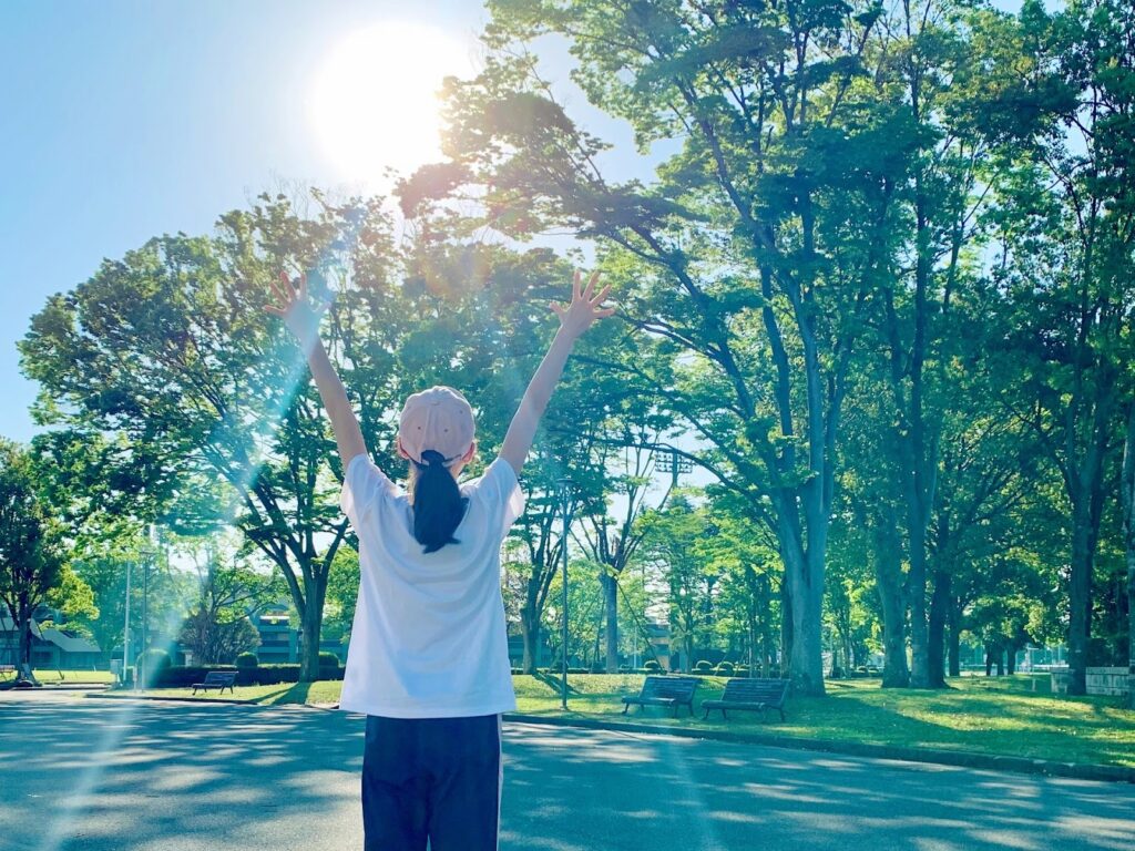 公園で両手を高く上げ、朝日を浴びている人の後ろ姿。木々の間から差し込む光が爽やかで、新しい一日の始まりを感じさせる：スピリチュアル・プラクティス