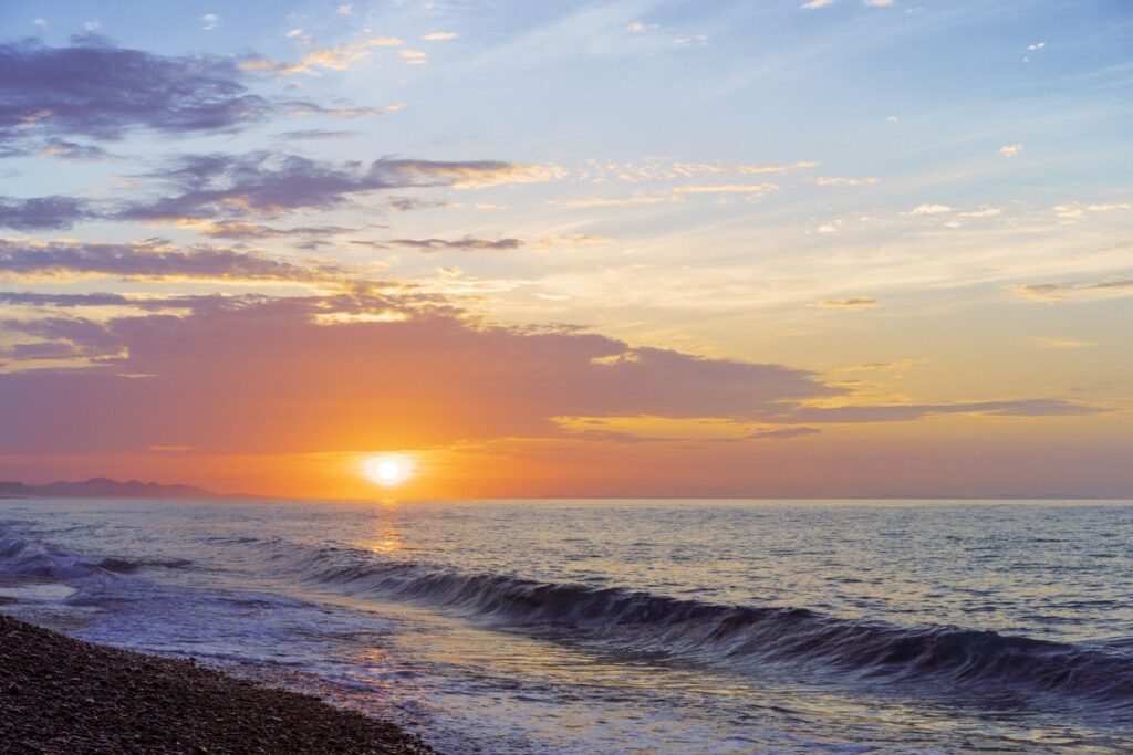 海辺から見た夕日。波が穏やかに寄せる中、オレンジと紫が混ざり合う美しい夕焼けが広がり、静かな時間を感じさせる風景：スピリチュアル・プラクティス
