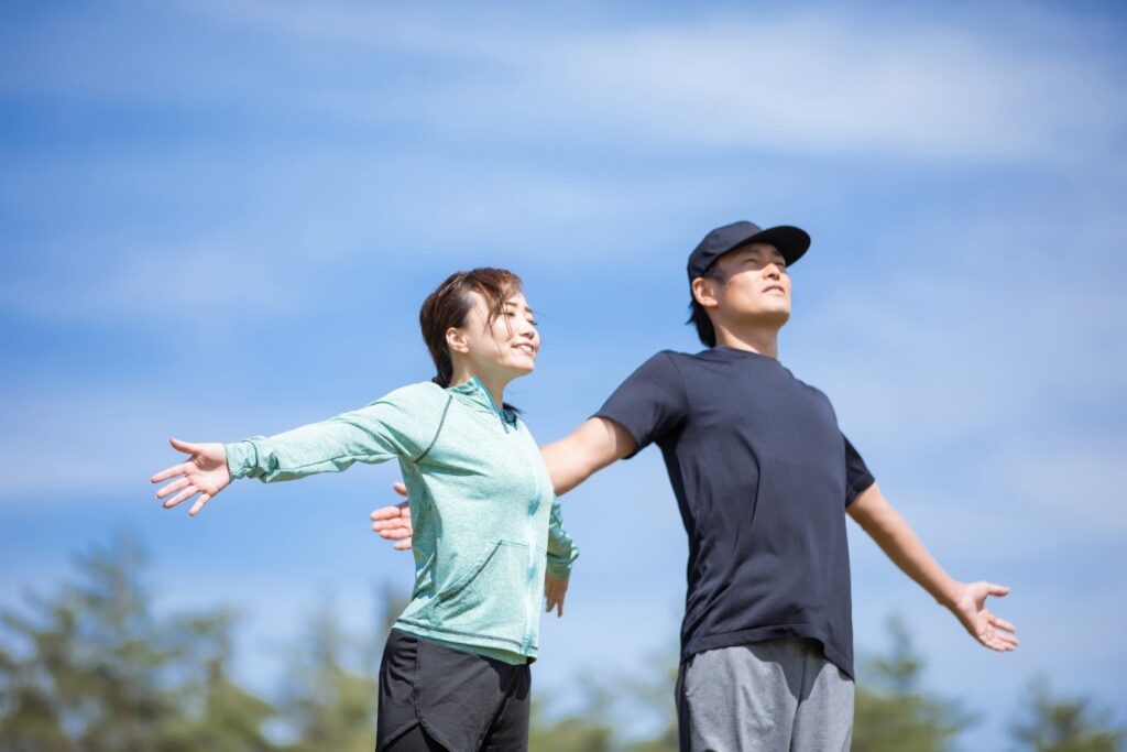 青空の下で両手を大きく広げ、深呼吸をする男女。自然の中で新鮮な空気を感じながら、心も体もリフレッシュしている様子:マインドフルネス・エクササイズ