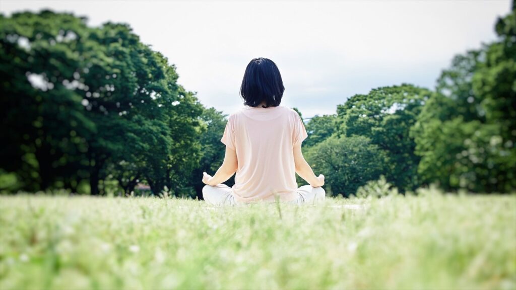 公園の芝生の上であぐらをかき、瞑想する女性の後ろ姿。緑に囲まれた静かな空間で、心を落ち着けるように深い呼吸をしている様子:マインドフルネス・エクササイズ