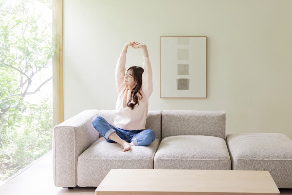 A woman sits on a bright living room sofa, stretching her arms and relaxing comfortably in the soft morning light: Meditation for Beginners