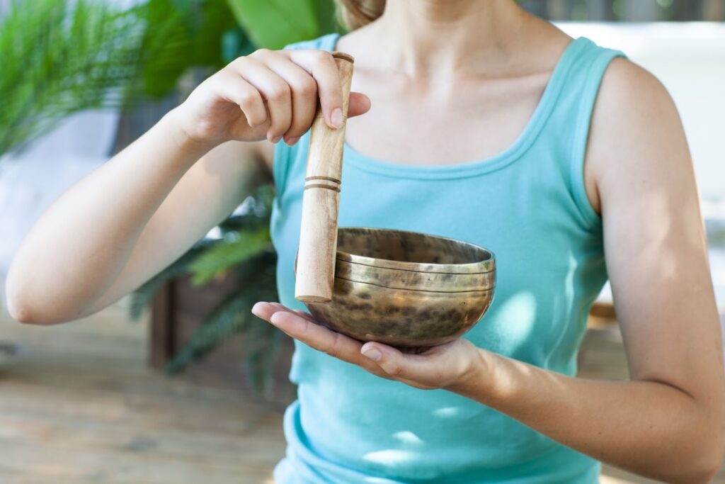 青いタンクトップを着た女性がシンギングボウルを手に持ち、木のスティックで縁を鳴らしている。穏やかな空間で瞑想やサウンドヒーリングを行っている様子:チベタン・シンギングボウル