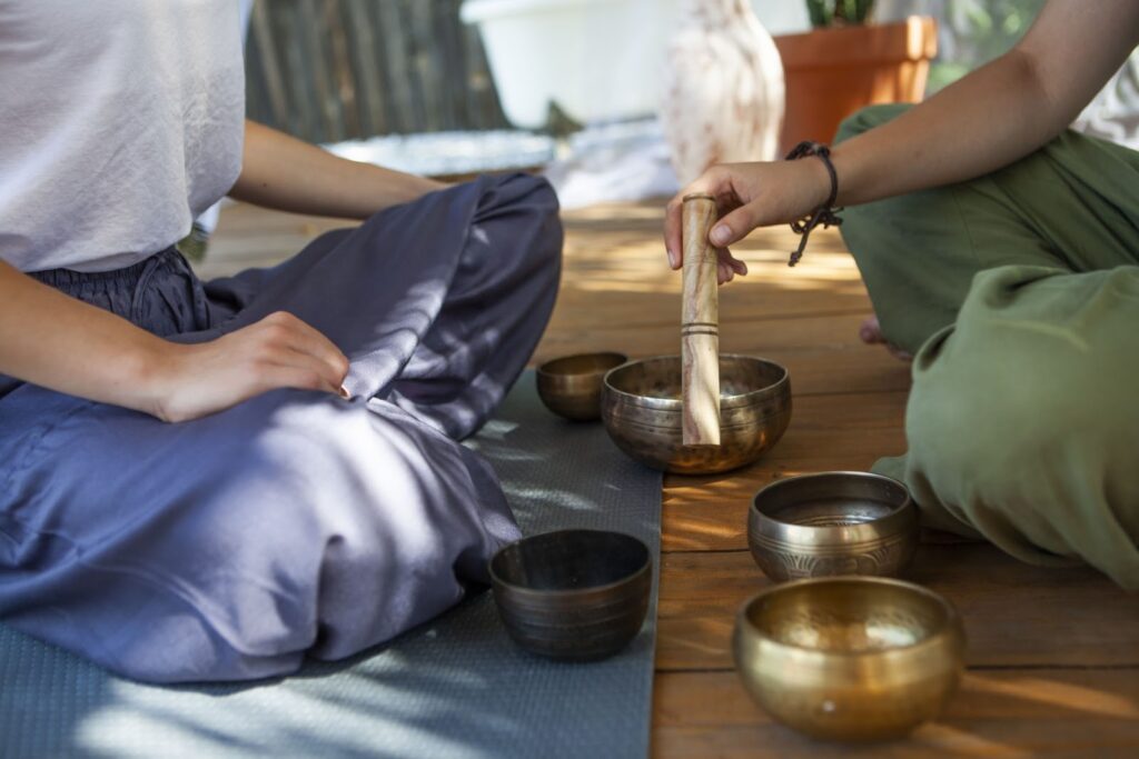 床に座って瞑想する2人の人。1人はシンギングボウルを木のスティックで鳴らしており、複数のボウルが前に並べられている。穏やかな音とリラクゼーションを感じさせる場面:チベタン・シンギングボウル