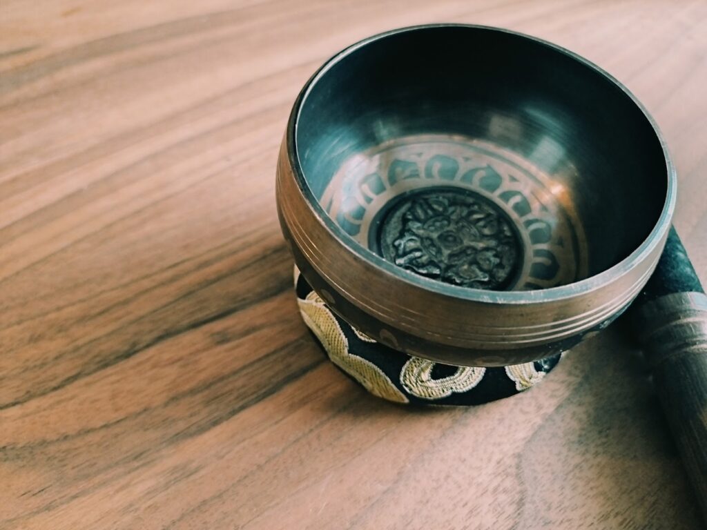 木製のテーブルの上に置かれたシンギングボウル。中央には装飾模様があり、横にはスティックが添えられている。瞑想やサウンドヒーリングを象徴するイメージ:チベタン・シンギングボウル