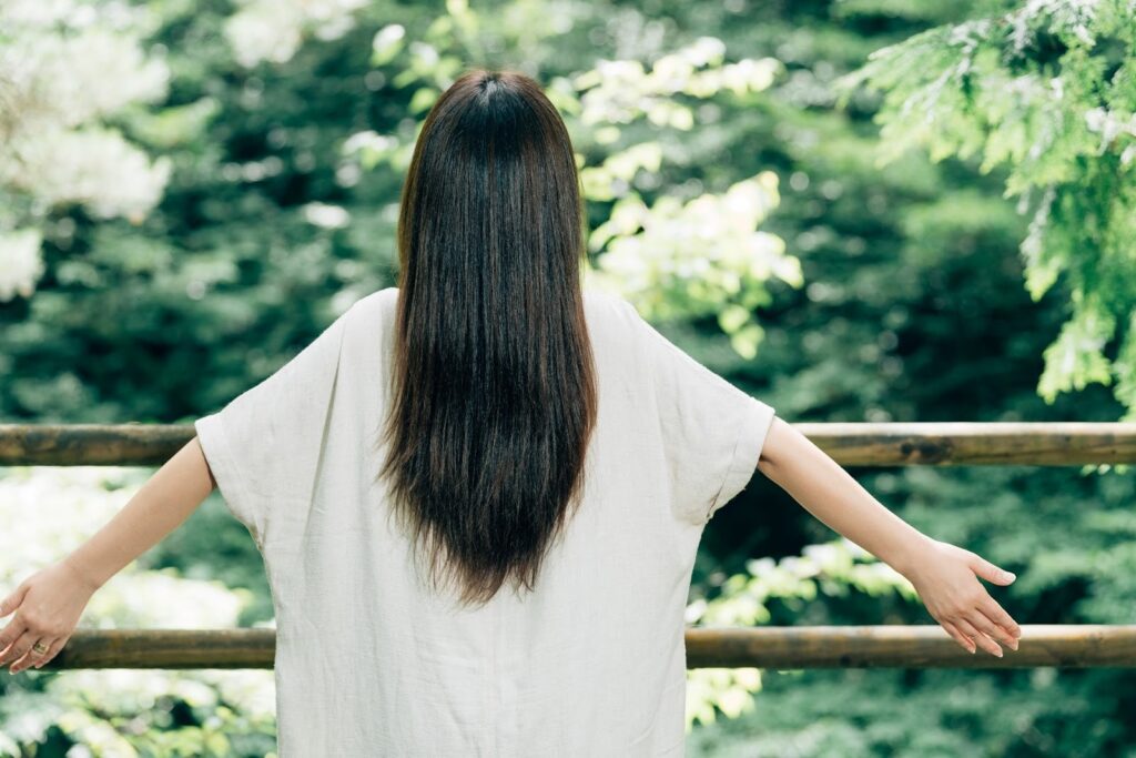 緑に囲まれた屋外で、白い服を着た女性が背中を向けて両腕を広げている姿。自然の中で深呼吸し、心を落ち着けているような印象のシーン:チベタン・シンギングボウル