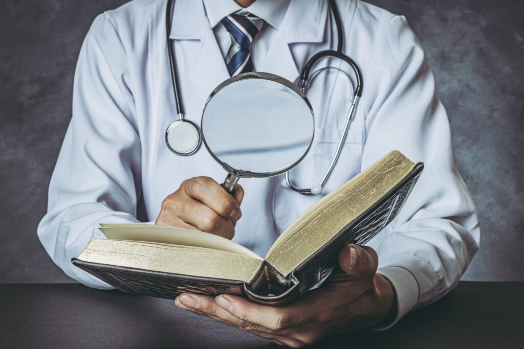 白衣を着た医師が聴診器を首にかけ、本を読みながら虫眼鏡でページを拡大して見ている様子。医療と研究を象徴するシーン:ジョン・カバット・ジン