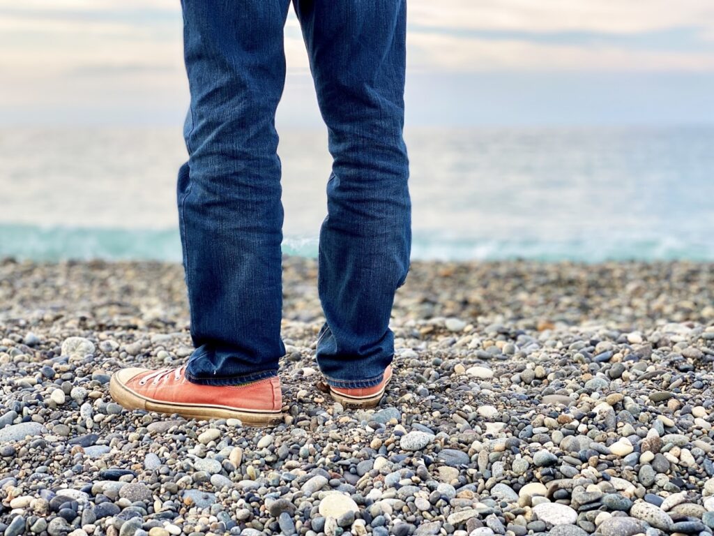 海辺の小石の上に立つ人の足元。青いジーンズとオレンジ色のスニーカーを履いており、背景には穏やかな海と空が広がっている:ジョン・カバット・ジン