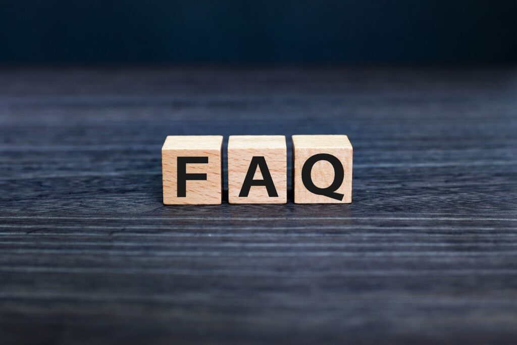 木製ブロックに「FAQ」と印字された文字が並び、暗い木目のテーブルの上に置かれている。背景はぼかされており、中央の「FAQ」が強調されている:ジョン・カバット・ジン