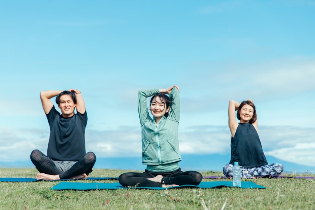 青空の下、芝生の上でヨガをする3人の男女。腕を上に伸ばしながら笑顔でストレッチをしており、爽やかで開放的な雰囲気：リラックス法
