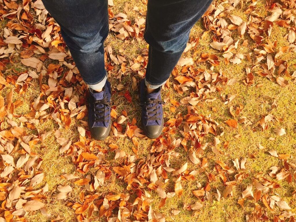 落ち葉が散らばる地面の上に立つ人物の足元。青いスニーカーとジーンズが秋の柔らかな光に照らされている。季節の移ろいを感じさせる風景：マインドフルネス・アクティビティ