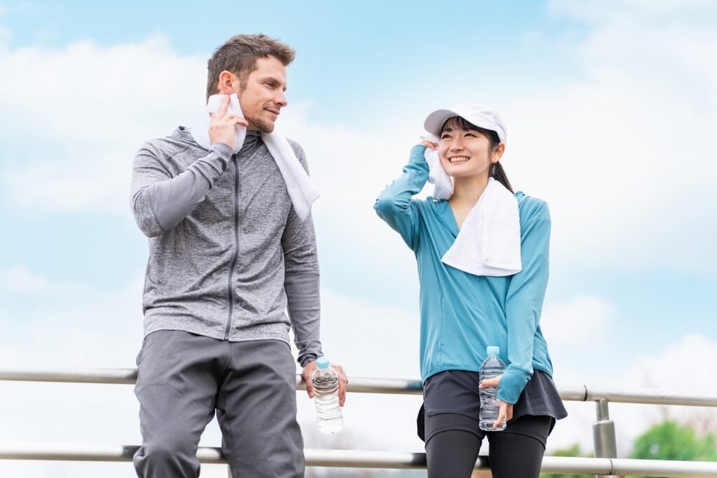 運動後に休憩しながら笑顔で会話する男女。タオルで汗を拭き、水のボトルを手にしている。青空の下で爽やかな雰囲気：マインドフルネス・アクティビティ