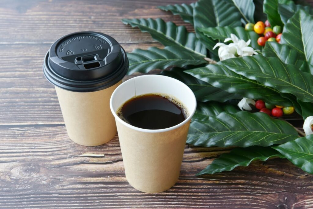 木製のテーブルの上に置かれた2つの紙コップ入りコーヒー。横にはコーヒーの葉と赤い実、白い花が添えられており、自然と香りを感じさせるイメージ：マインドフルネス・アクティビティ