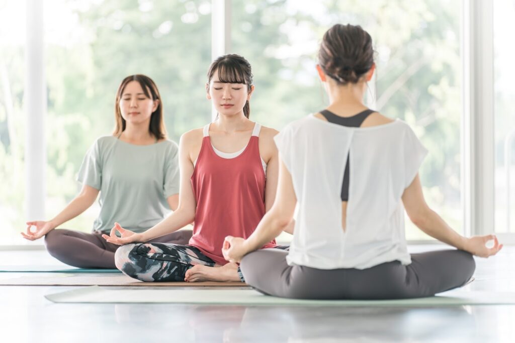 Three women sitting on yoga mats with their eyes closed in meditation, in a bright, peaceful room filled with natural light: Spiritual Retreat