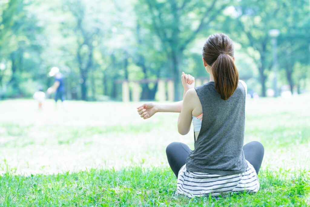 A woman stretching on a grassy field under bright sunlight, seen from behind, creating a calm and relaxed atmosphere. Trees and a parent with a child are softly blurred in the background: Spiritual Retreat