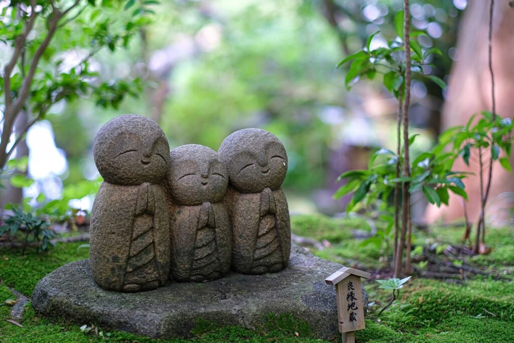 Three small stone Jizo statues standing on moss-covered ground, hands folded in prayer, surrounded by lush greenery, creating a peaceful and serene atmosphere: Spiritual Retreat