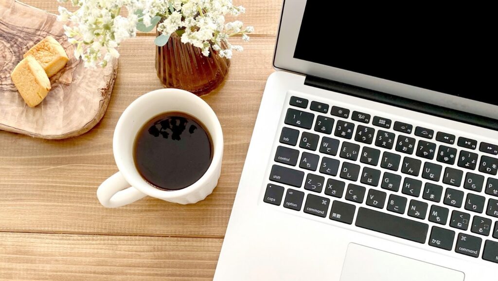 A laptop, cup of coffee, cookies, and a small vase with flowers on a wooden table, evoking a relaxed atmosphere of remote work or a cozy workspace: MBSR