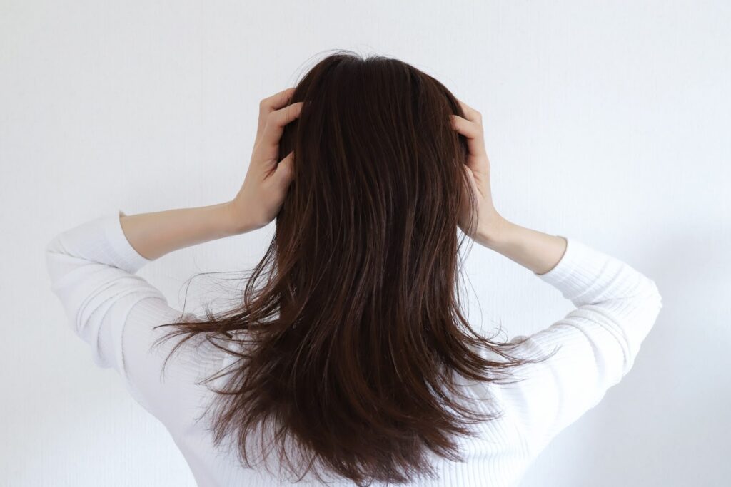 白い服を着た人が後ろを向き、両手で頭を抱えている様子。髪が肩にかかり、ストレスや悩みを感じているような印象の構図：リラックス法