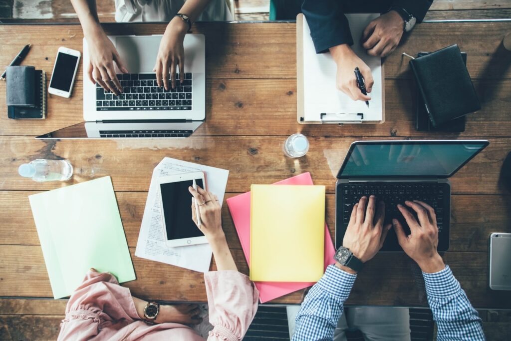 Close-up of several people working together at a wooden table with laptops, tablets, and documents, representing teamwork and collaboration in a meeting or office setting: MBSR