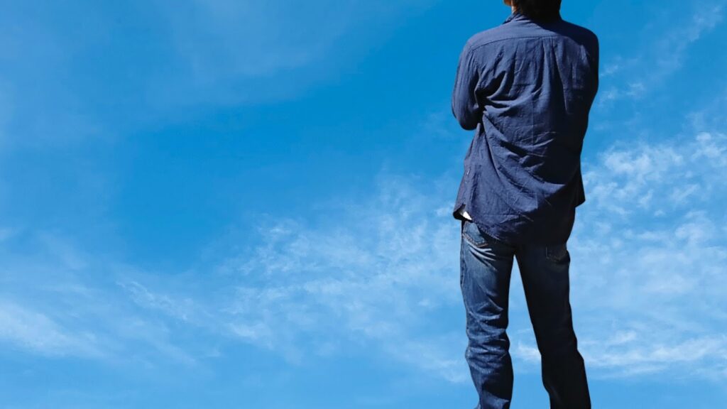 A person wearing jeans and a shirt standing with their back to the camera under a bright blue sky, evoking a sense of freedom and openness: MBSR