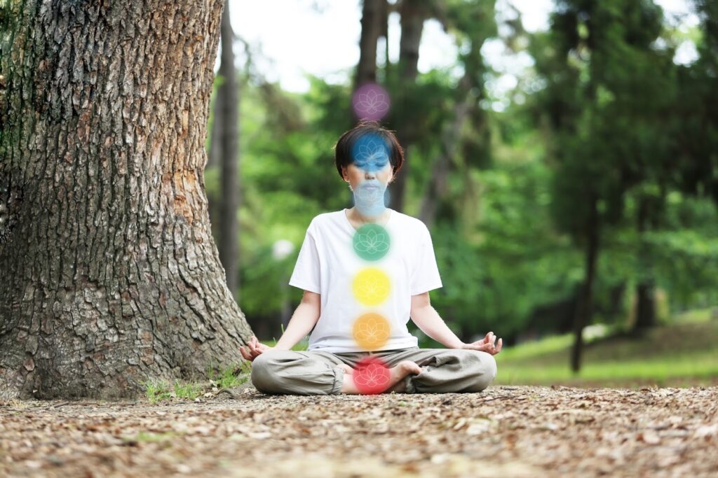 大きな木のそばで座禅を組み、瞑想している人。体の中心には赤・黄・緑・青・紫のチャクラを象徴する光が並び、心身の調和を表している:座蒲