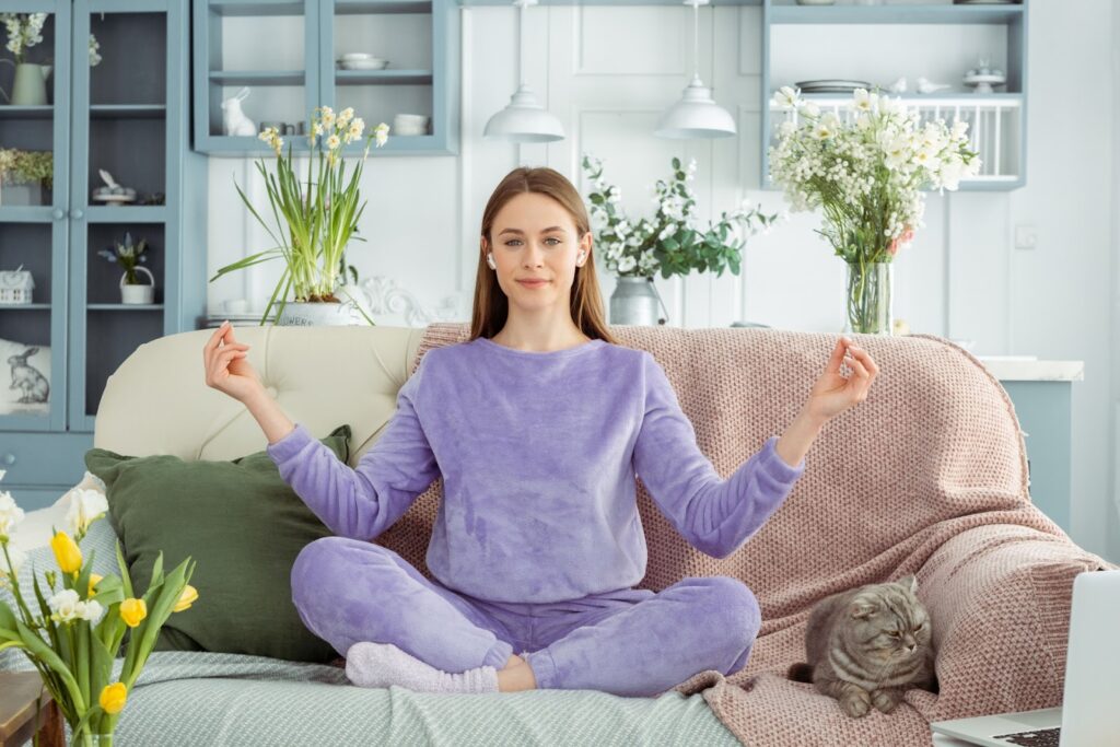 リビングのソファで瞑想する女性。紫のルームウェアを着てイヤフォンをつけ、隣には穏やかにくつろぐ猫がいる。花が飾られた明るい室内の雰囲気:座蒲