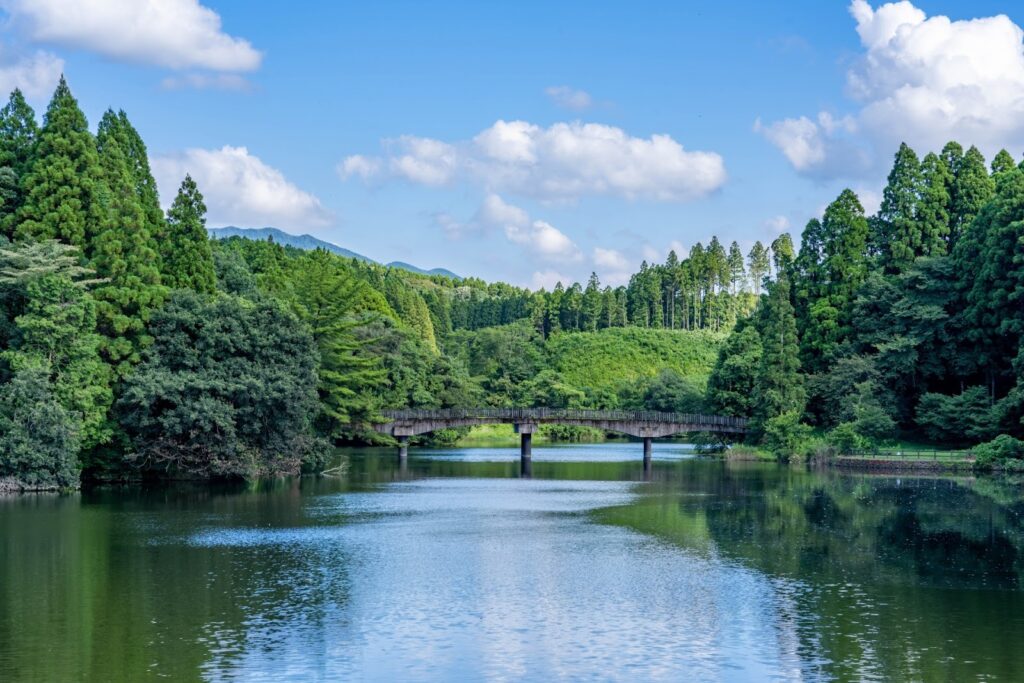 静かな湖の上に木製の橋がかかり、周囲を青々とした森と山々が取り囲んでいる。澄んだ空と白い雲が水面に映り込む美しい自然風景：静けさの瞑想