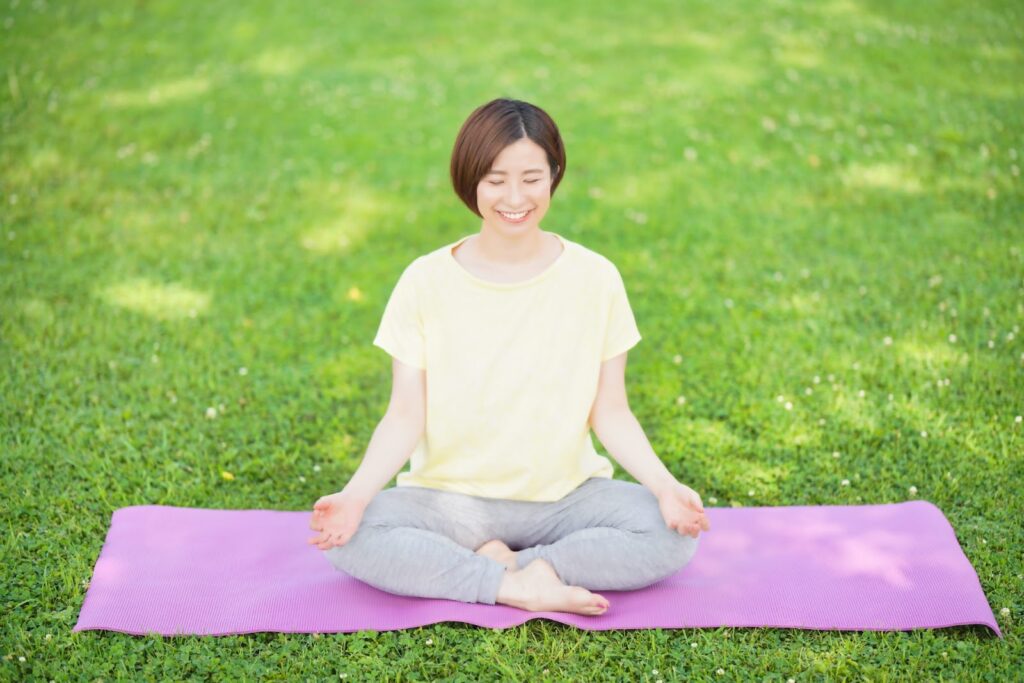 芝生の上でヨガマットに座り、目を閉じて穏やかに瞑想する女性。自然の中でリラックスしながら心を落ち着けている様子：静けさの瞑想