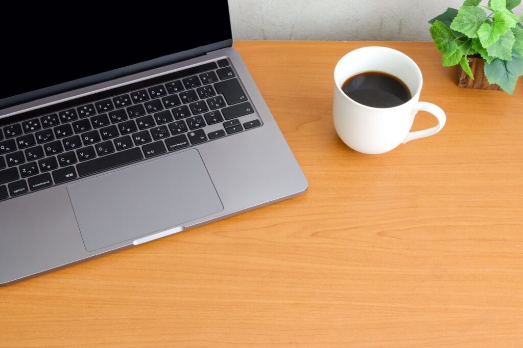 木製のデスクの上にノートパソコンと白いコーヒーカップ、そして小さな観葉植物が置かれている。シンプルで落ち着いた作業スペースの様子：静けさの瞑想