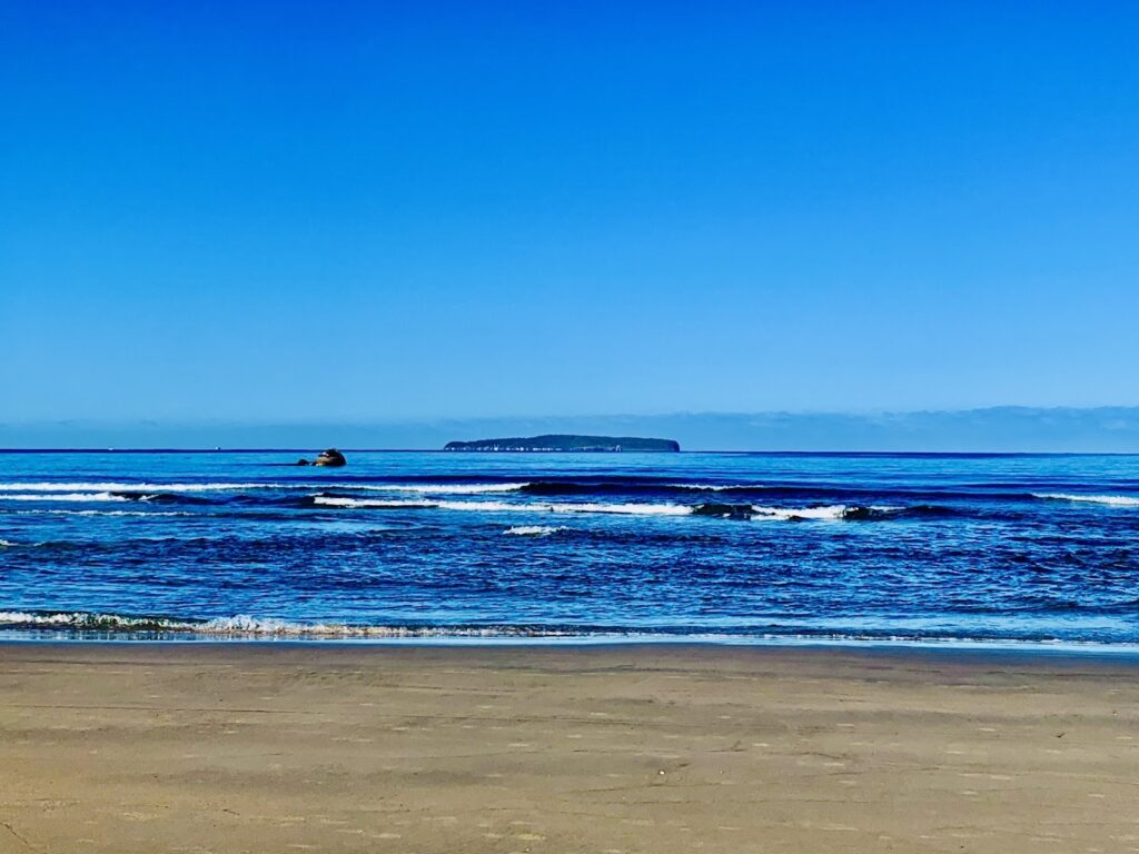 青い空と穏やかな海が広がる風景。遠くに小さな島が見え、静かな波が砂浜に寄せている。静寂と広がりを感じさせる美しい海岸の光景：TM瞑想